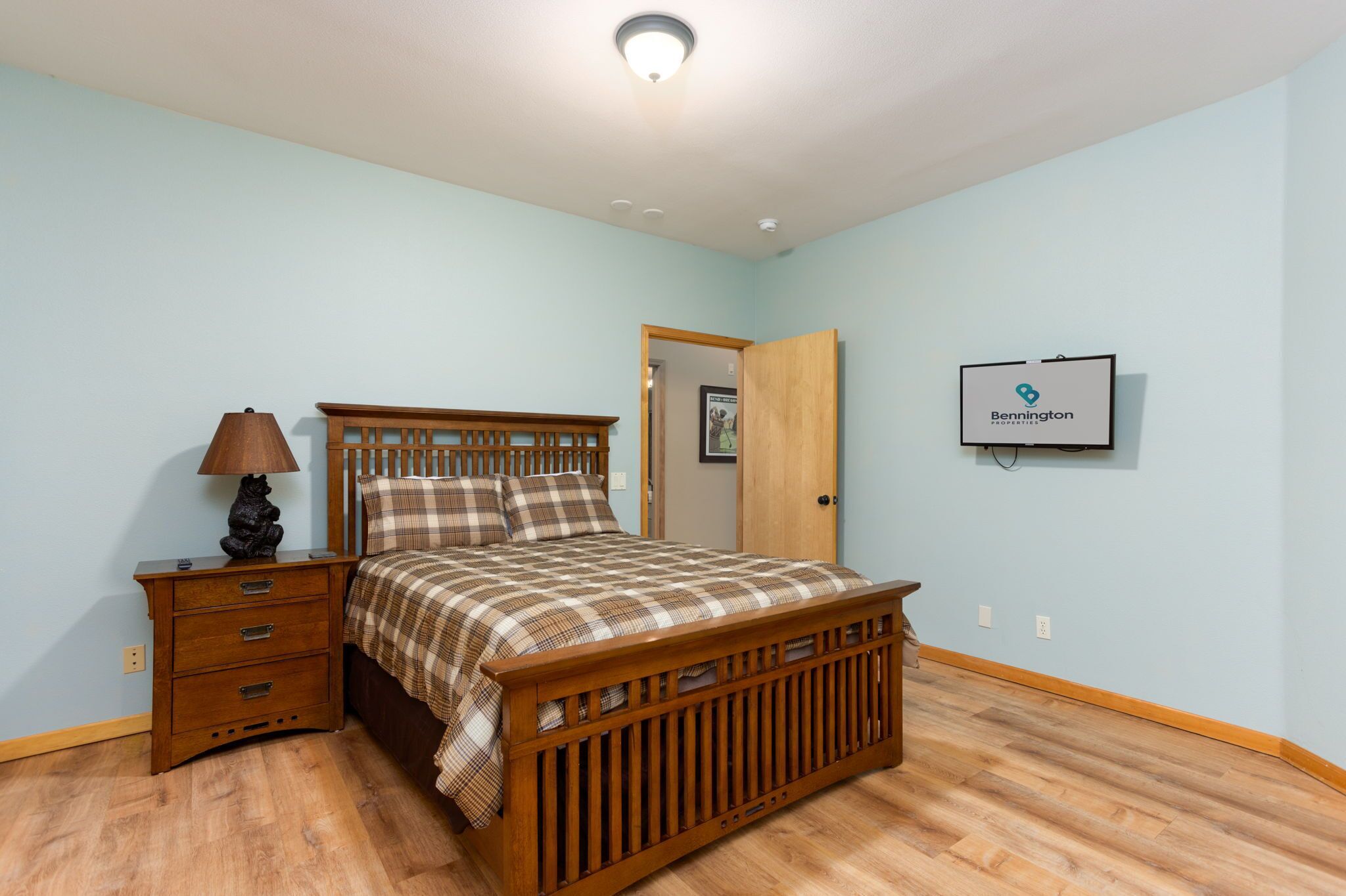 A bedroom with a wooden bed, a nightstand with drawers, a lamp, a framed picture, and a sign on the wall.