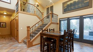 A spacious dining area with a wooden table and chairs, a staircase with a wooden railing, and a view through large glass doors. - Cypress 4 - Huge Home, Foosball, BBQ, Dog Friendly (Sunriver)
