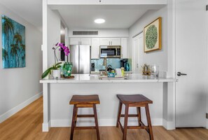 A modern kitchen with white cabinetry, a stainless steel refrigerator, a microwave, and a coffee maker. There are two wooden stools at the counter.