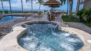 A hot tub with a view of the ocean, lounge chairs, and a thatched umbrella.
