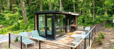 Una cabaña moderna con cerramiento de vidrio, terraza de madera y zona de estar al aire libre rodeada de bosque.