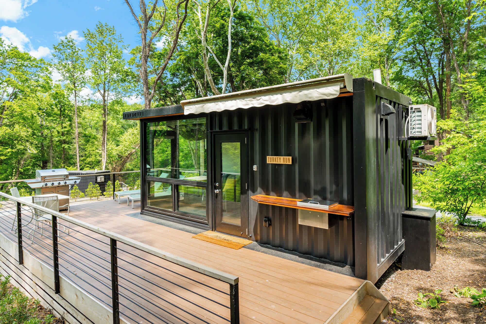 Een klein, modern bouwwerk buiten met een glazen deur, een houten terras en een bord waarop 