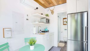 Una cocina compacta con refrigerador de acero inoxidable, microondas y estufa pequeña. Hay un lavamanos con grifo, una planta en maceta sobre la encimera y una silla azul verdosa junto a una mesa pequeña.