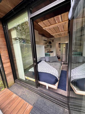 A modern bedroom with a large bed, a small kitchenette, and a sliding glass door leading to a balcony. - Cute Riverside Cabin w Hot Tub, Fire Pit, & Kayaks (Luray)