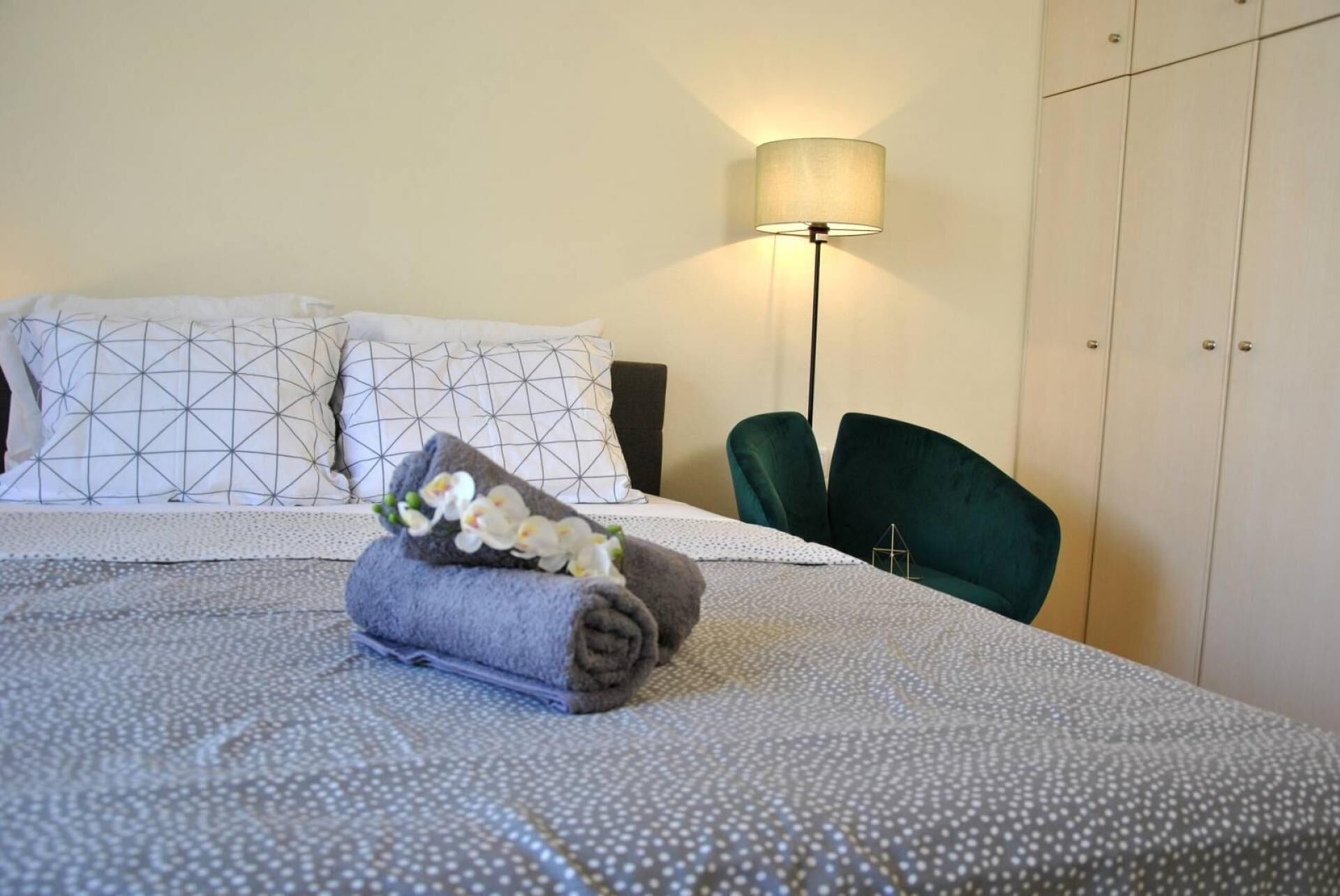 Ein ordentlich bezogenes Bett mit gerolltem Handtuch und Orchideen, eine Stehlampe und ein grüner Sessel in einem Hotelzimmer.