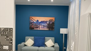 A modern living room with a blue accent wall, a sofa with patterned cushions, a painting of a cityscape, and a dining area with a table and chairs. - The Comfortable House 2 (Genova)