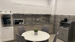 A modern kitchen with white cabinets, a built-in oven, microwave, and gas stove. There is a round dining table with chairs and a potted plant. - The Comfortable House 2 (Genova)