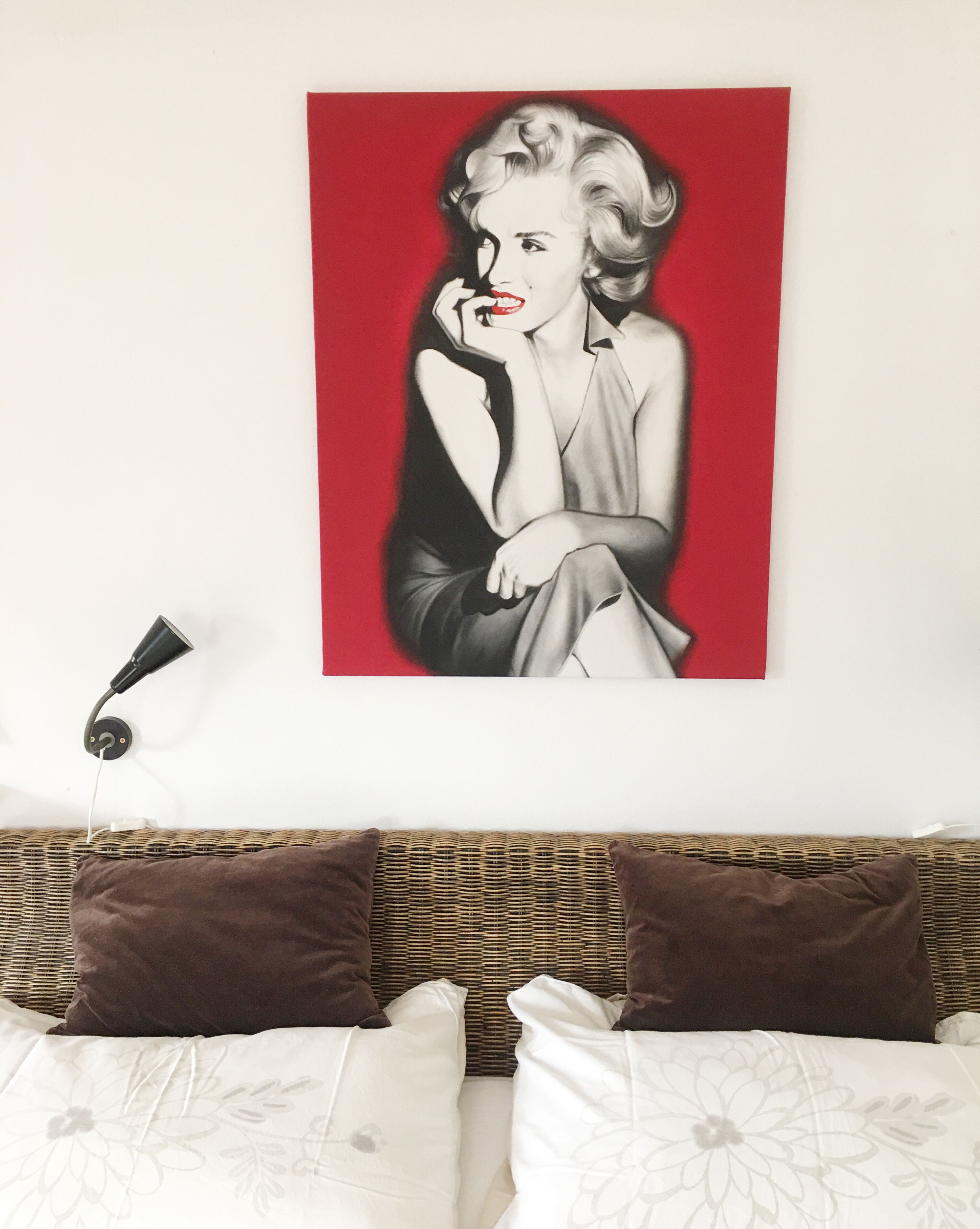 A room with a bed, two brown pillows, a white floral bedspread, and a red and black artwork on the wall.