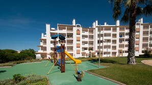 A playground with a slide and swings, surrounded by a well-maintained lawn and palm trees, with a multi-story building in the background. - Ondas Verdes - Central, Pool & Tenis Court (Albufeira)