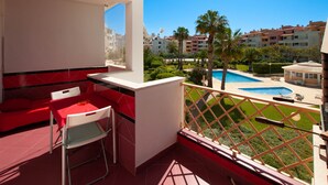 A balcony with a red sofa, a small table, and a chair. There is a pool with a palm tree and a building in the background. - Ondas Verdes - Central, Pool & Tenis Court (Albufeira)