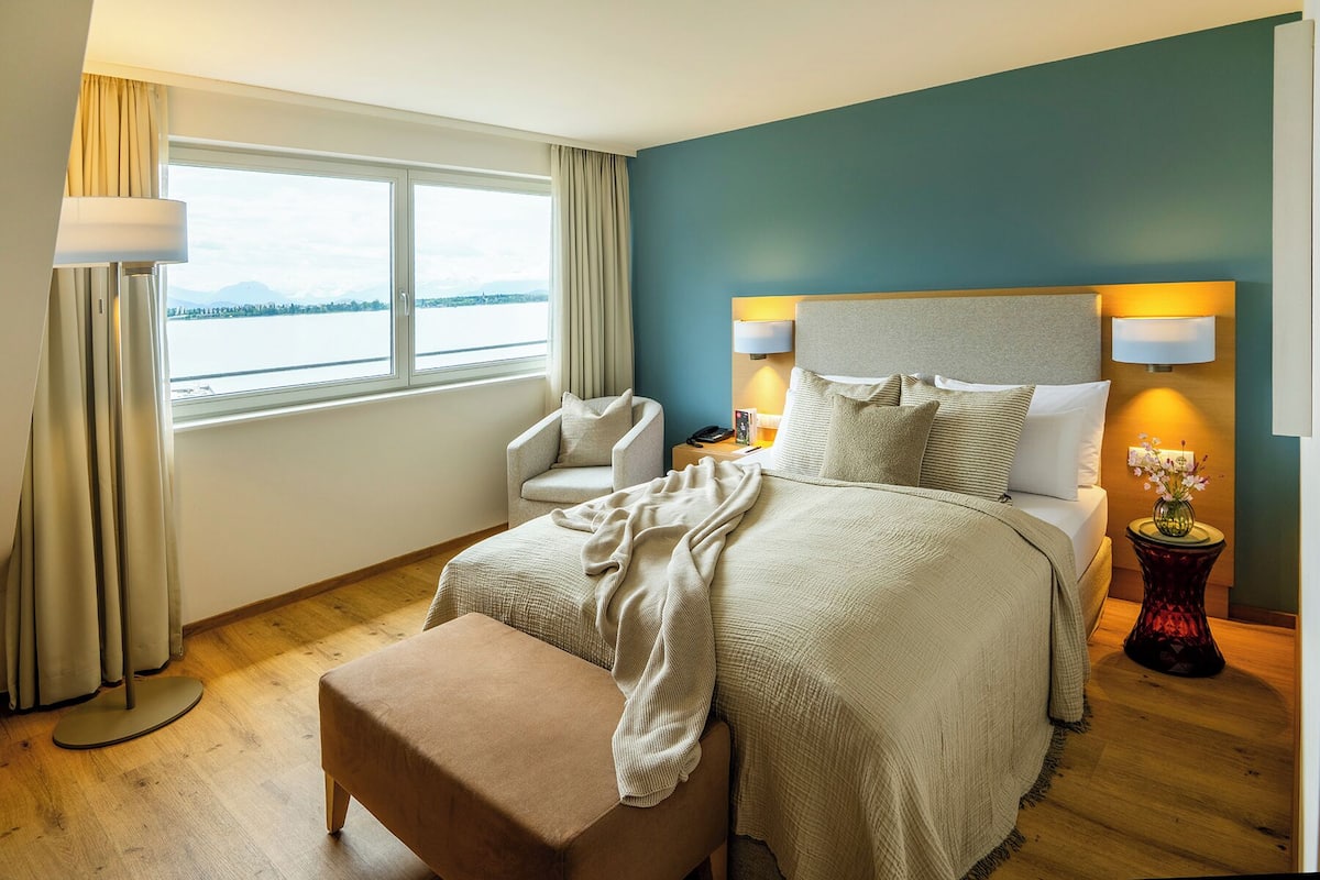 A hotel room with a bed, a chair, a nightstand, a lamp, and a view of a body of water through a window.