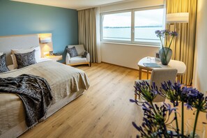 Habitación de hotel con cama, una silla, una mesita y ventana con vista al agua.
