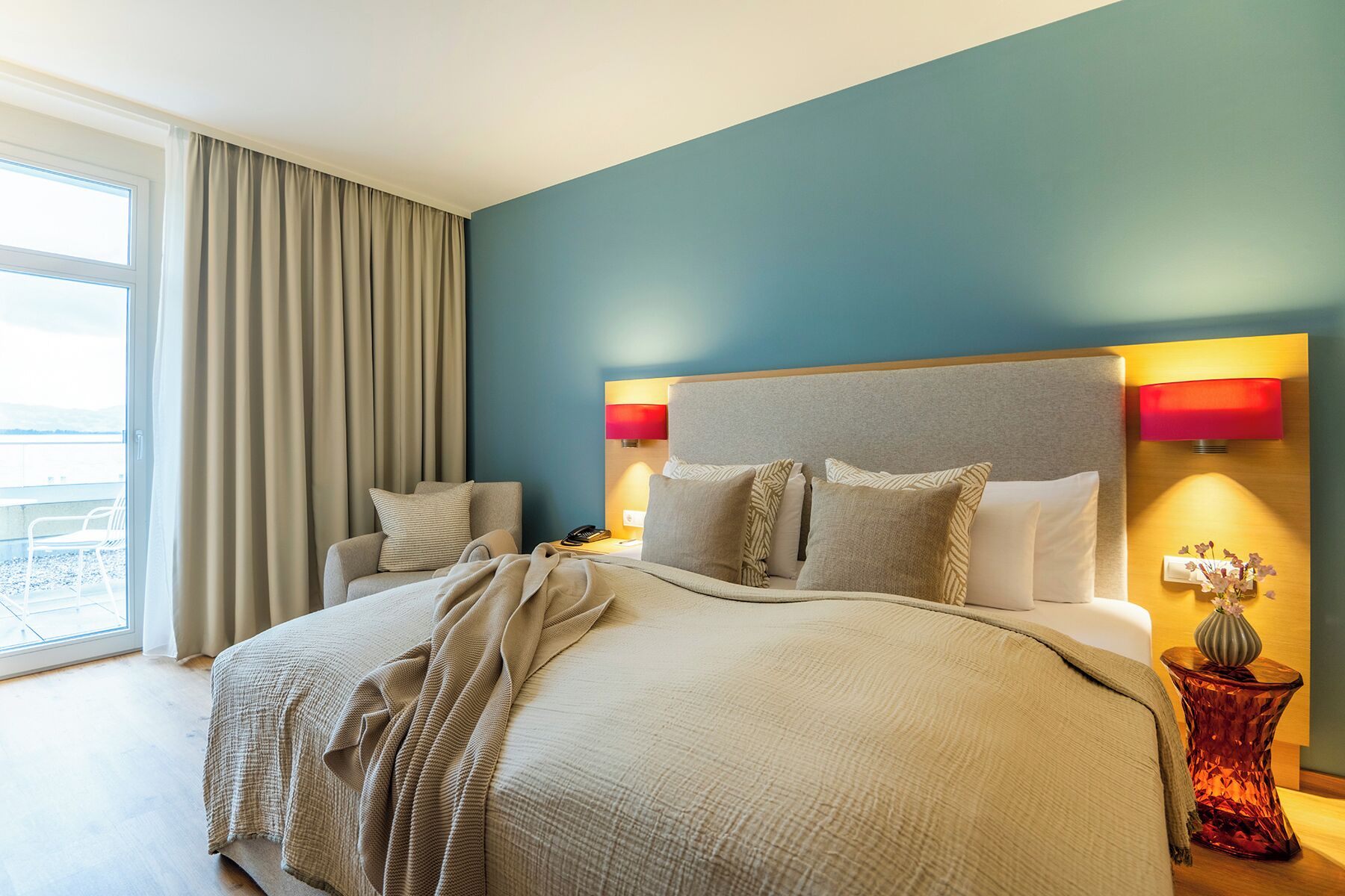 Hotelkamer met een groot bed, een fauteuil, een nachtkastje, een vaas met bloemen en uitzicht op het landschap.