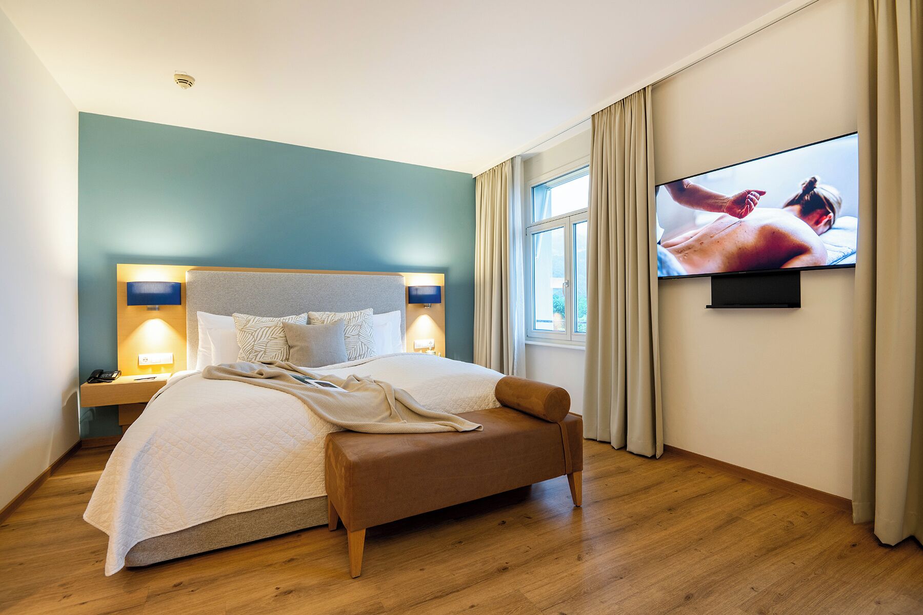Ein modernes Hotelzimmer mit einem großen Bett, einem an der Wand montierten Fernseher und einem Nachttisch mit Lampe.