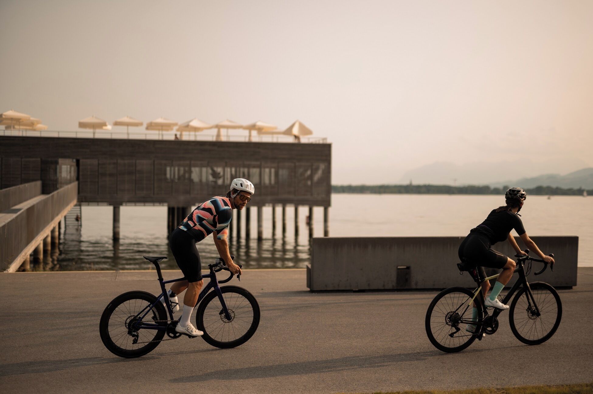 Two cyclists on a paved path with a waterfront and a pier in the background.