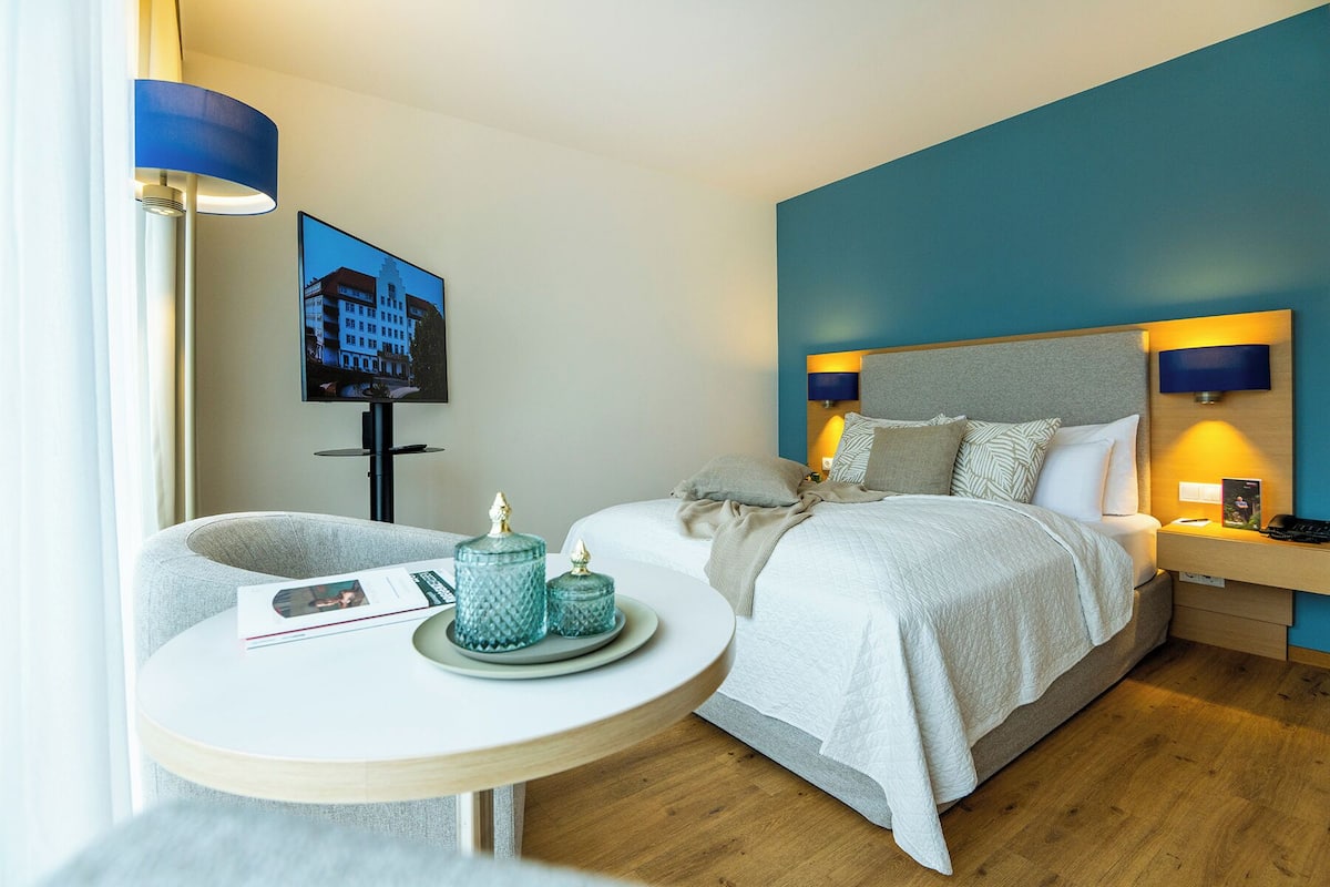 A hotel room with a bed, bedside lamps, a TV, a small table with a magazine, and decorative items.