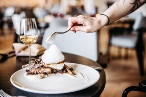 Un postre en un plato, un tenedor, una copa de vino blanco y una persona con un tatuaje en el brazo.