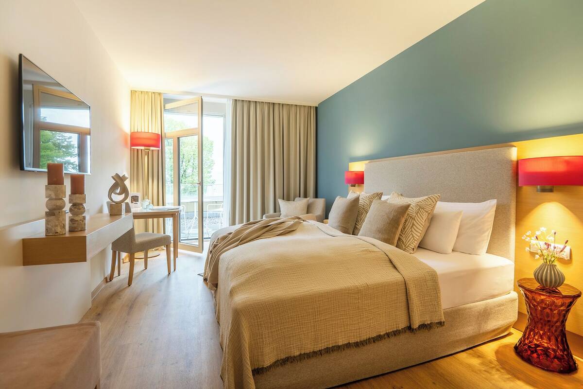 A modern hotel room with a large bed, a desk, and a chair. There is a balcony with curtains and a view of greenery.