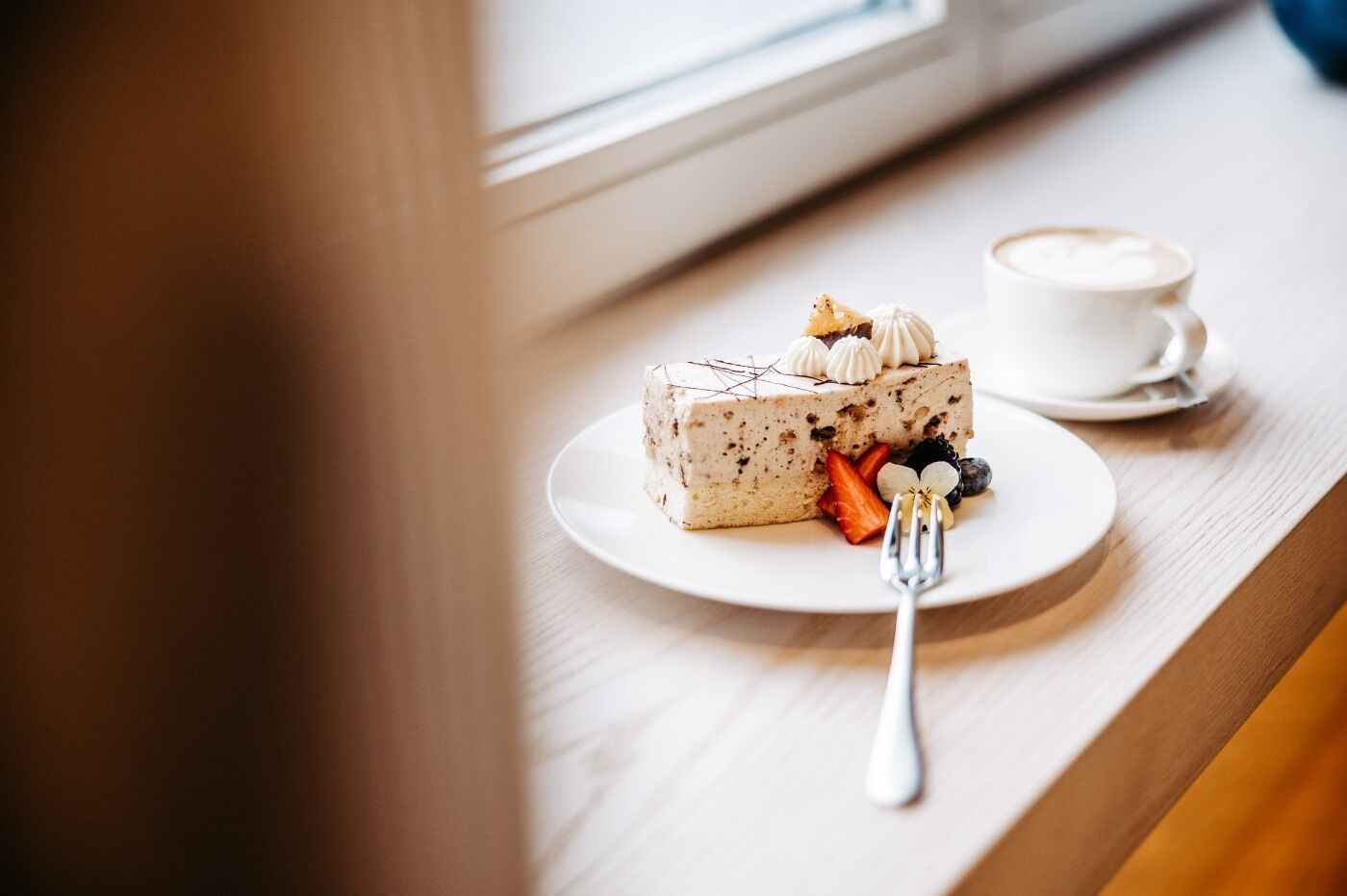A slice of cake with fruit on a plate, a cup of coffee, and a fork on a wooden table by a window.