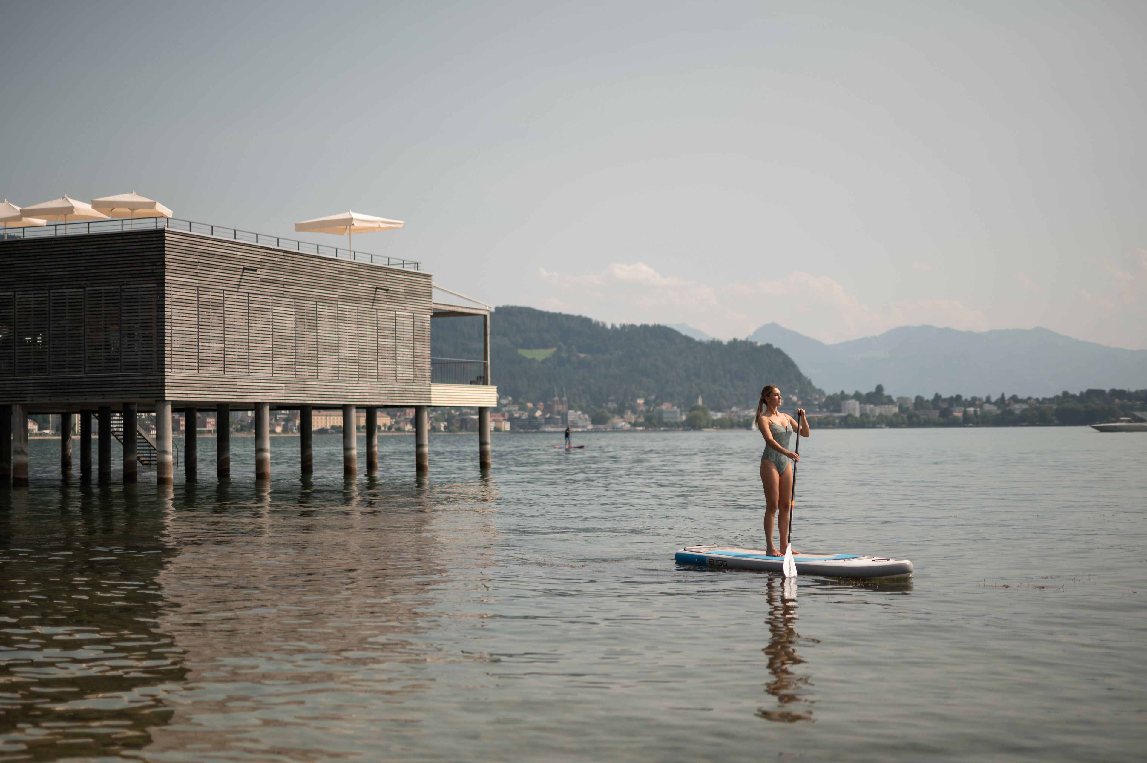 Eine Frau steht auf einem Stand-Up-Paddleboard auf einem ruhigen See, im Hintergrund ein Holzsteg und Sonnenschirme.