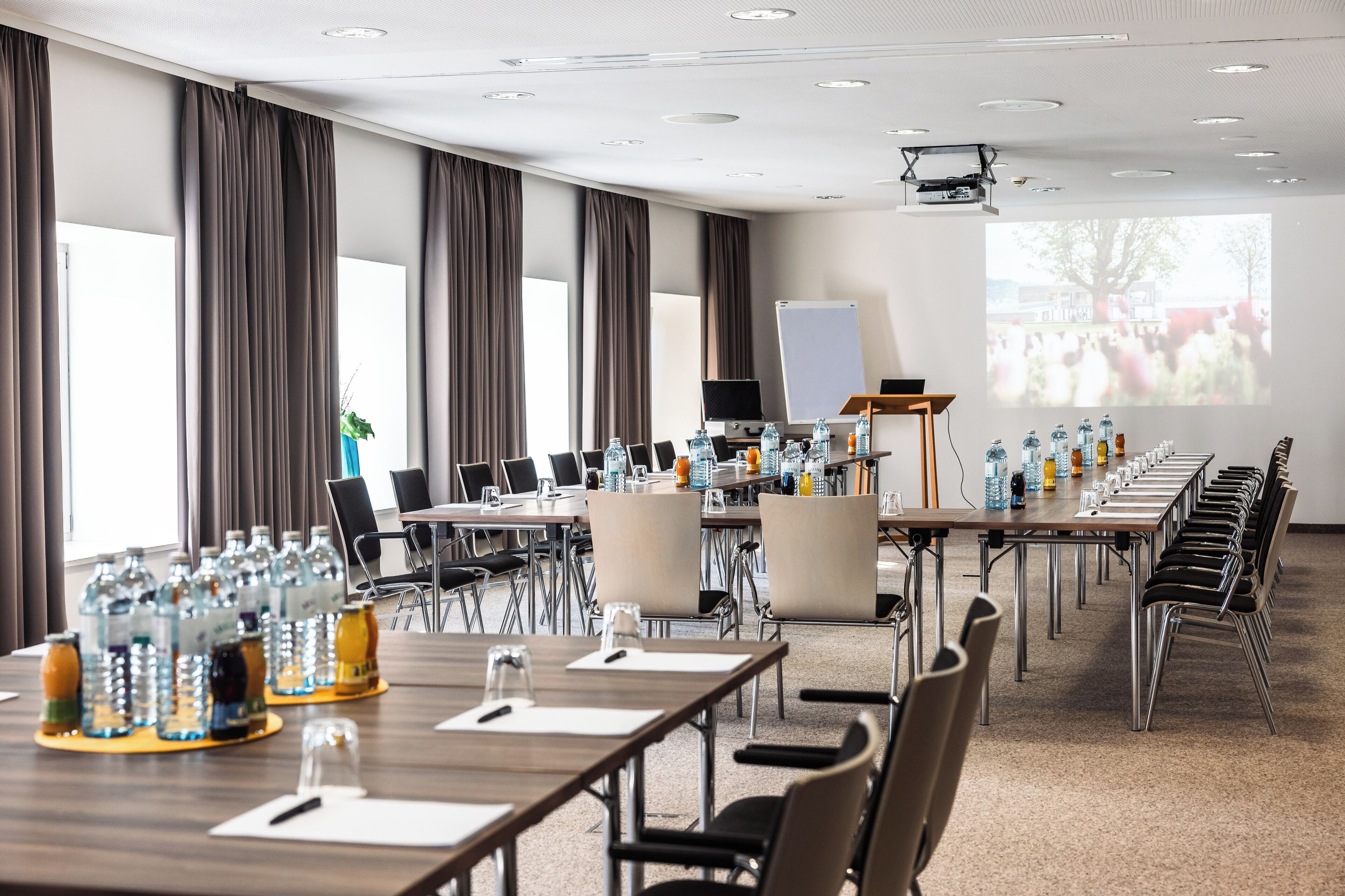 Un salón de conferencias con mesas largas, sillas, botellas de agua y una pantalla para proyector.