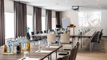 Un salón de conferencias con mesas largas, sillas, botellas de agua y una pantalla para proyector.