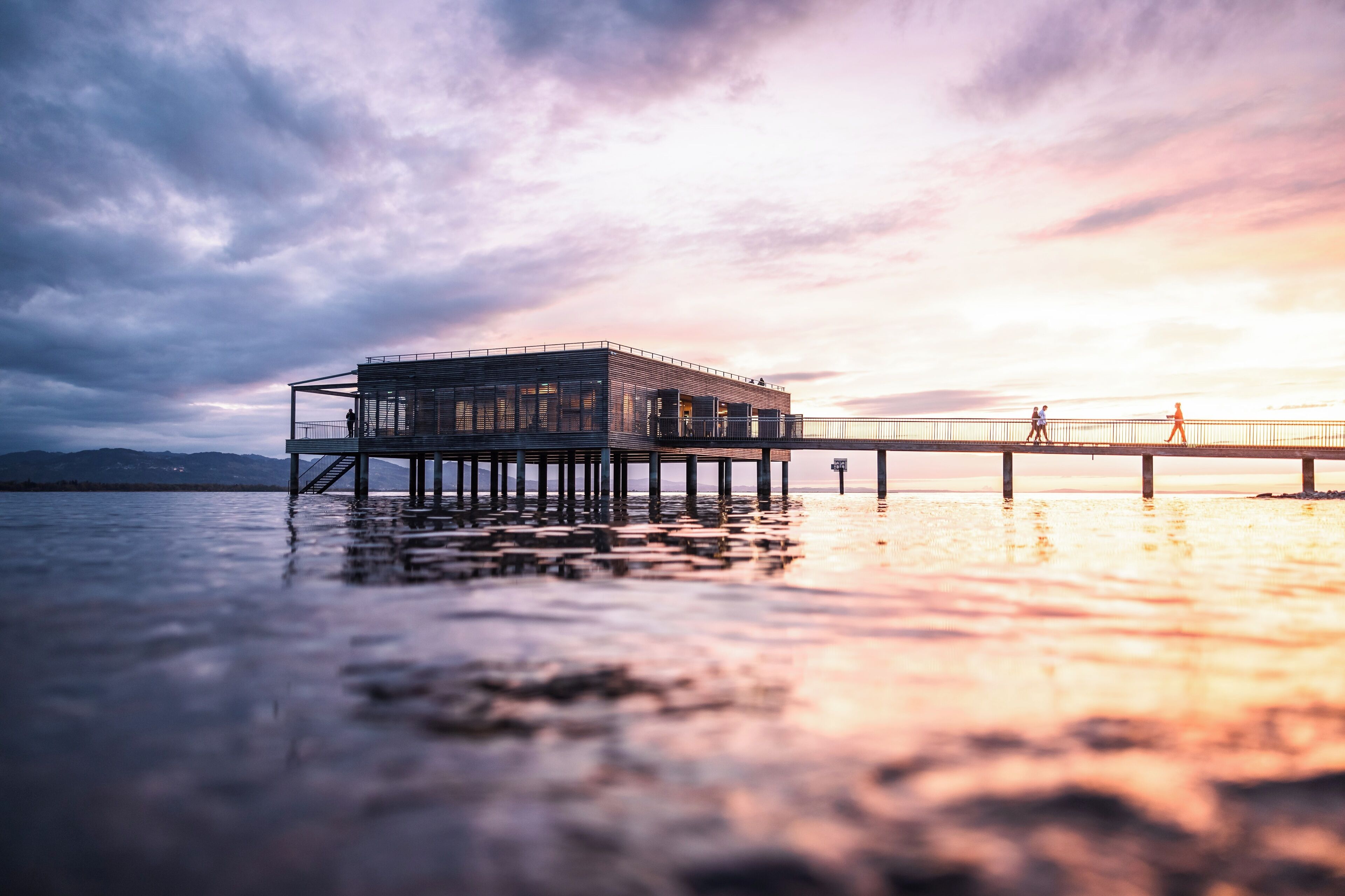 A modern building with a glass facade on stilts over water, a dock extending into the water, and a sunset in the background.