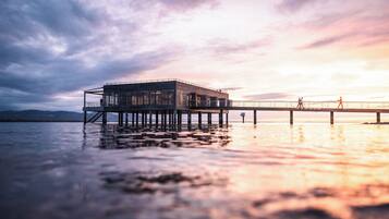 Ein modernes Gebäude mit Glasfassade auf Stelzen über dem Wasser, eine Anlegestelle, die sich ins Wasser erstreckt, und ein Sonnenuntergang im Hintergrund.