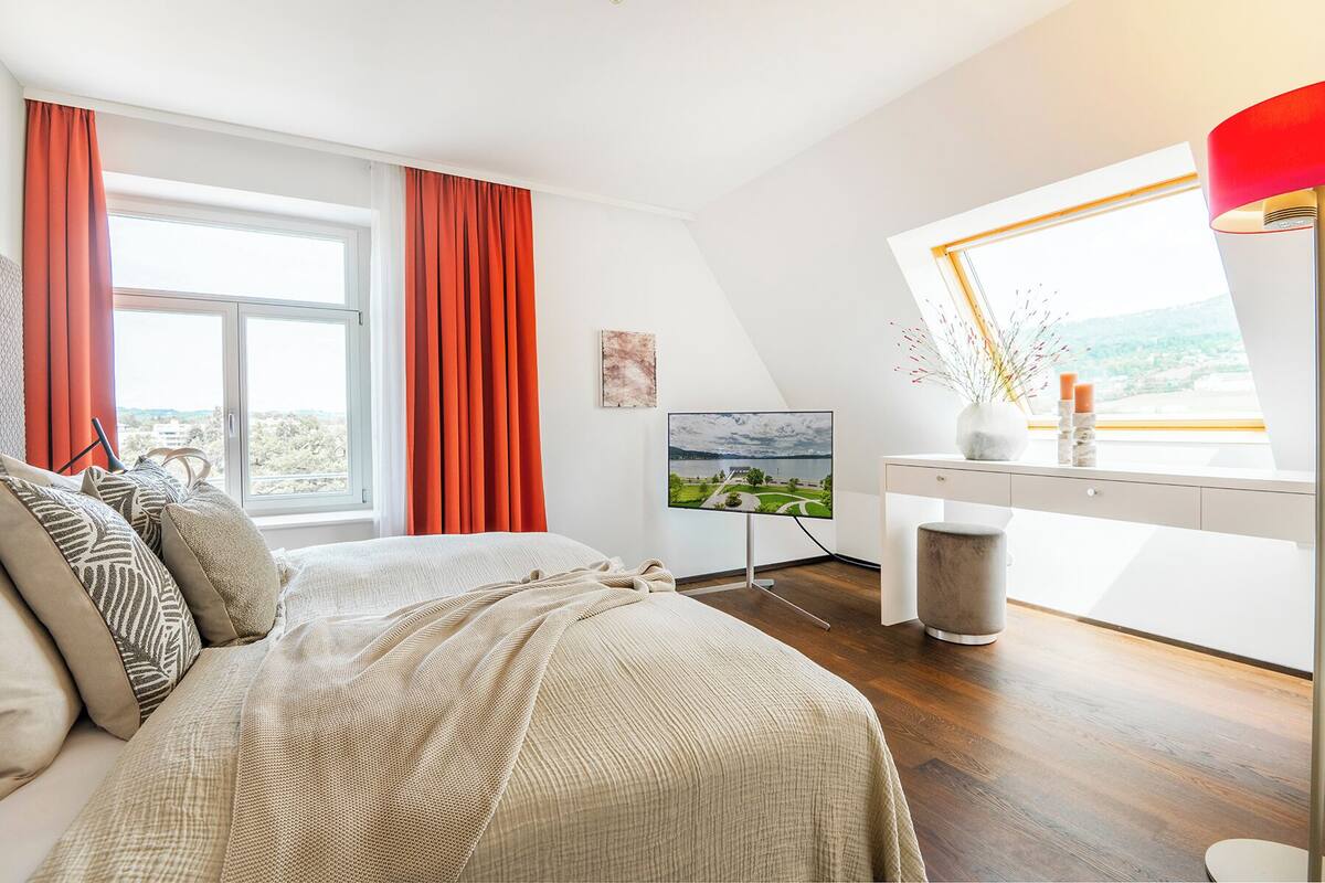 A modern bedroom with a bed, a desk, a TV, and a window with a view.