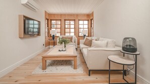 En moderne stue med trægulv, beige sofa, sofabord, fjernsyn monteret på væggen og et stort vindue med naturligt lys.