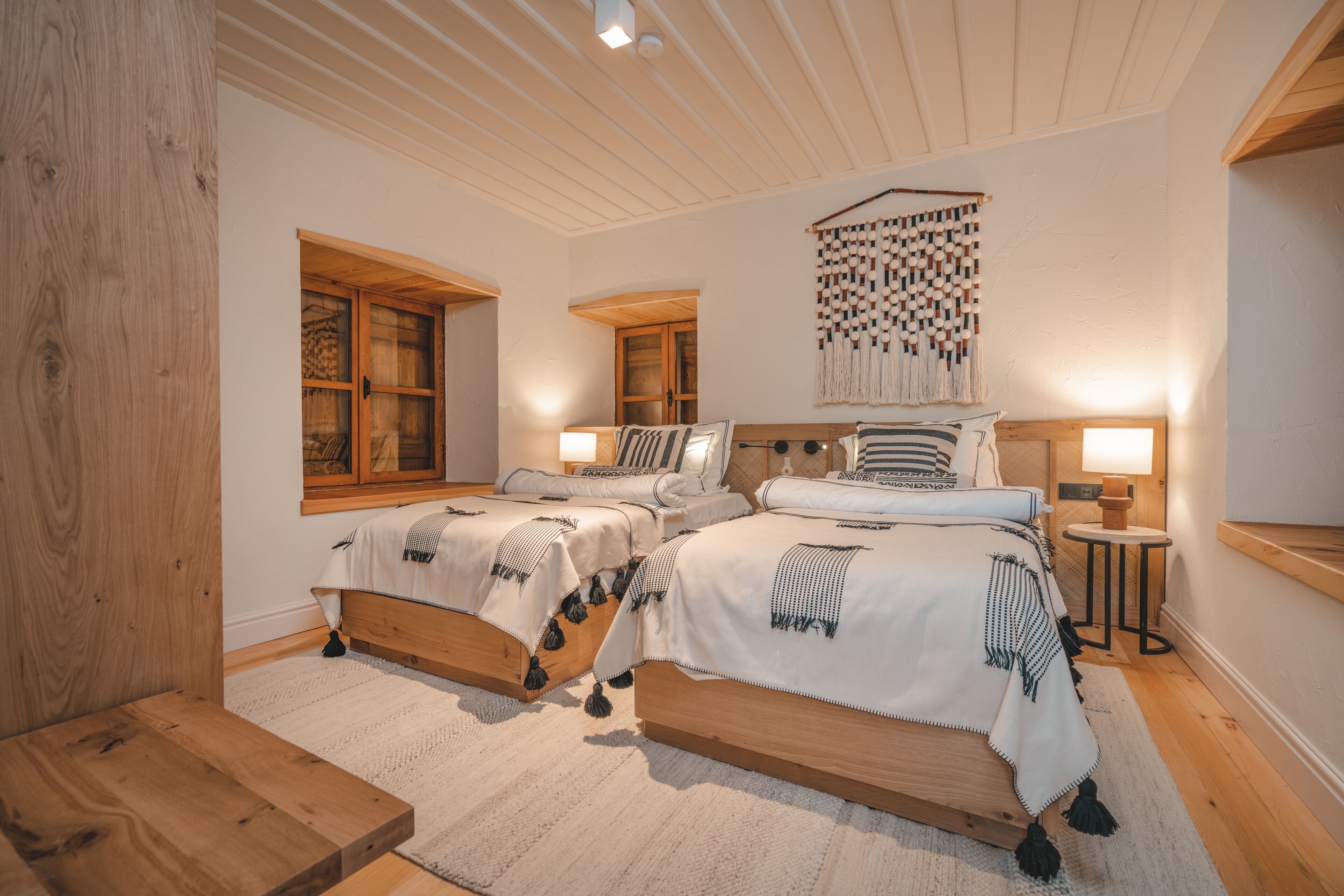 Un dormitorio con dos camas de madera, un ventanal grande, un tapiz en la pared y una mesita de noche con una lámpara.