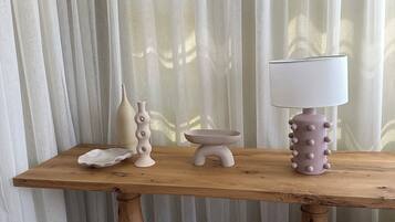 A wooden table with a white lamp, a decorative bowl, and a vase against a backdrop of sheer curtains.
