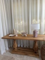 Una mesa de madera con una lámpara blanca, un cuenco decorativo y un florero frente a cortinas traslúcidas.