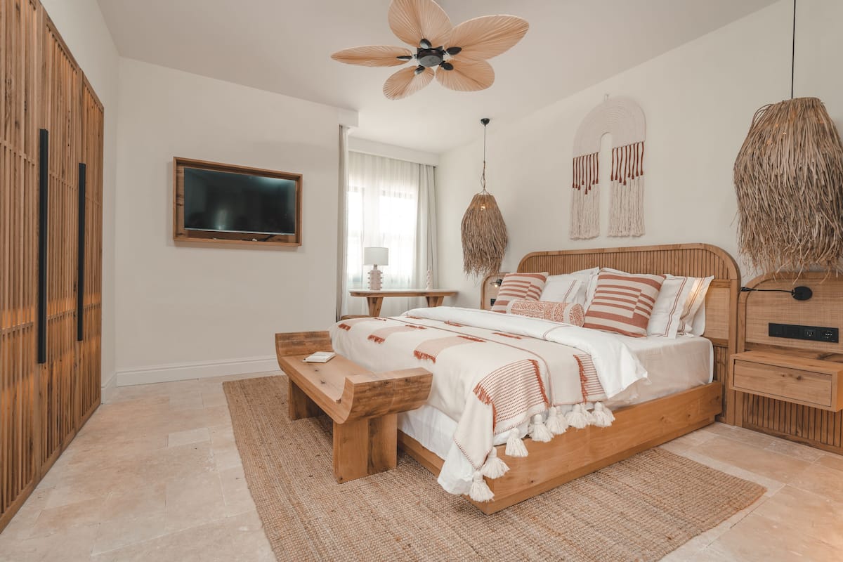 A bedroom with a wooden bed, a wooden bench, a ceiling fan, a TV, and a woven lamp.