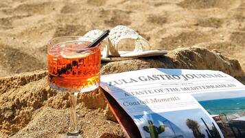 A beach scene with a glass of orange liquid, a straw, a straw hat, and a Zula Gastro Journal magazine on the sand.