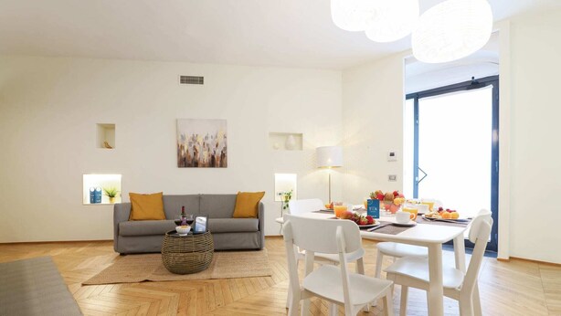 A modern living room with a grey sofa, a dining table with white chairs, and a large window. - Sweett | Maison Laghetto (Milano)