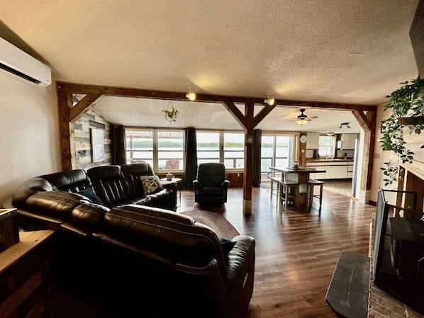 A spacious living area with a leather sofa, a dining table, and chairs, wooden beams on the ceiling, and large windows offering a view of the outdoors.