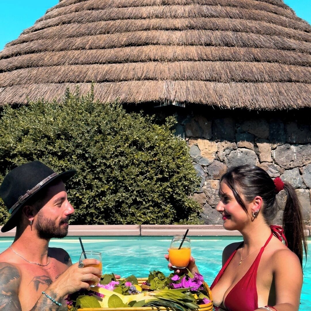 Un área de piscina con una estructura de techo de paja, una pared de piedra y abundante vegetación.