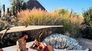 Una pareja descansando en una hamaca en un jardín con una estructura de techo de paja al fondo.