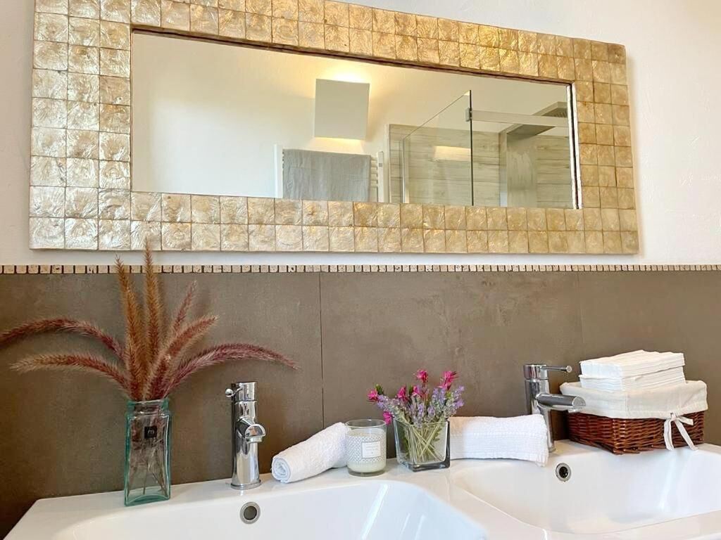 A bathroom with two sinks, a mirror with a textured frame, a vase with dried plants, and a basket of towels.