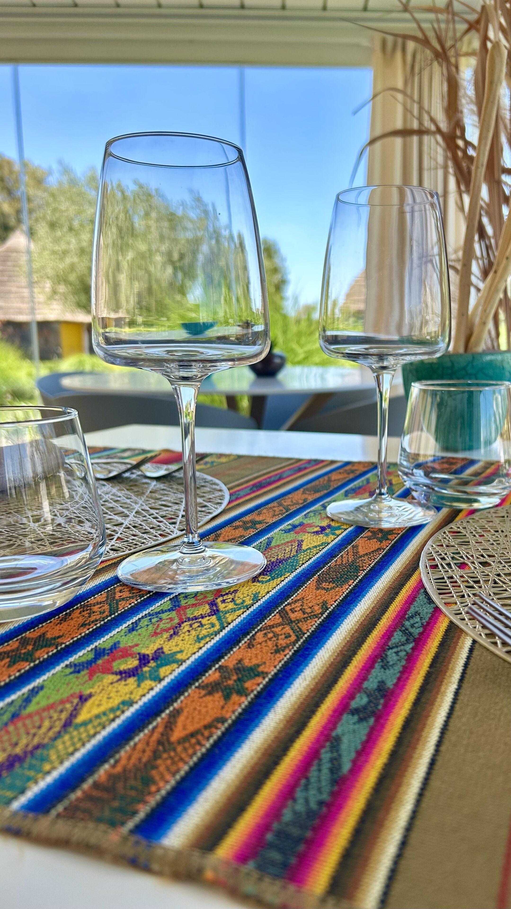 Two empty wine glasses on a colorful woven tablecloth with a view of greenery and a building in the background.