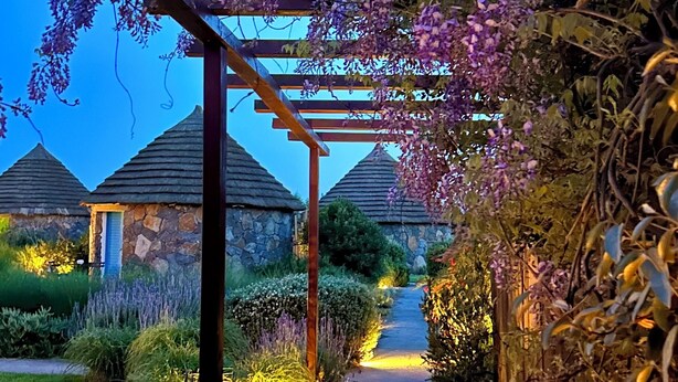 Un sendero bajo una pérgola adornada con flores moradas, que conduce a edificios de piedra con techos de paja.