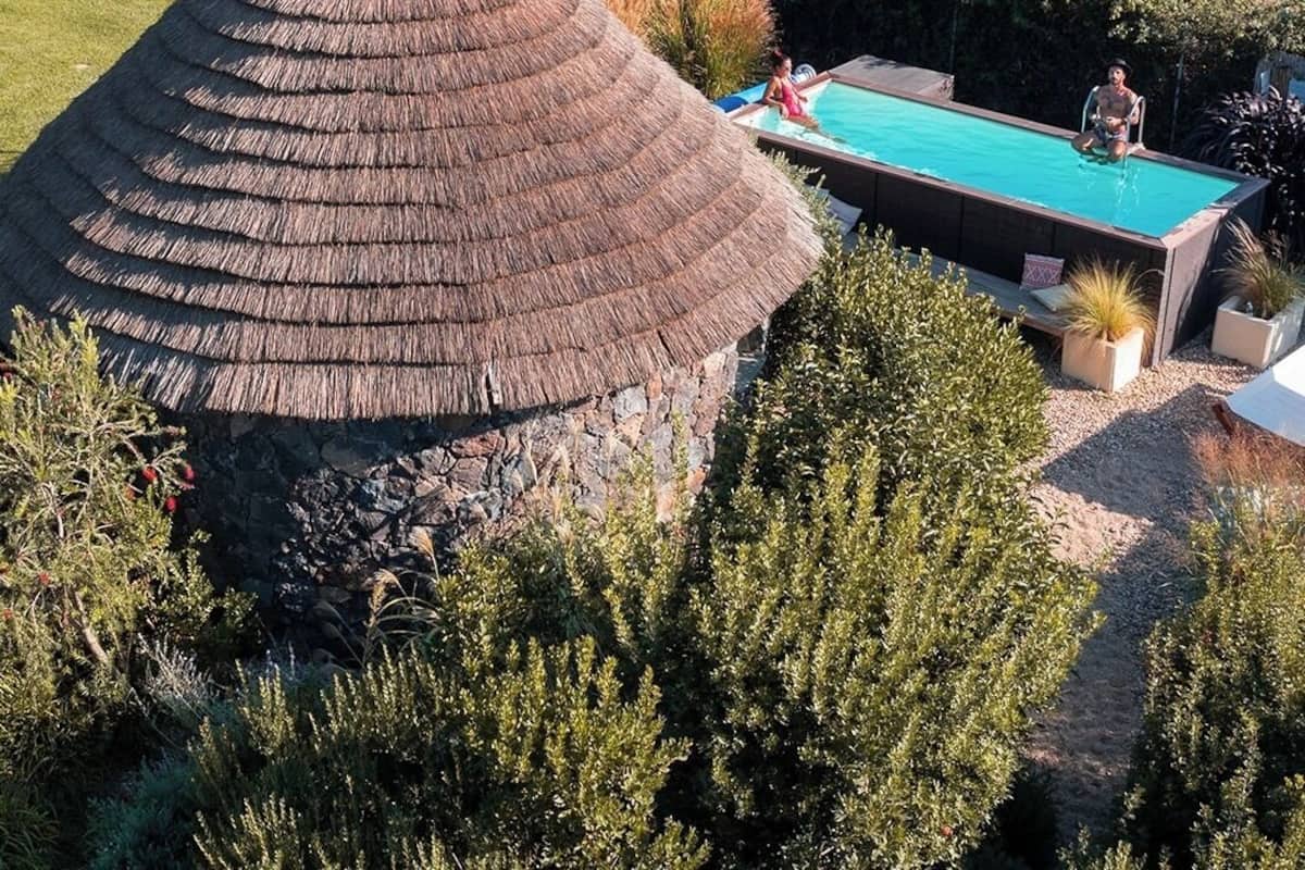 A thatched-roof structure with a swimming pool and a stone building in the background.