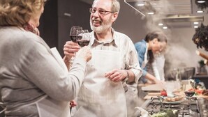Una persona con delantal blanco de pie en una cocina con estufa y ollas, rodeada de gente y una mesa de comedor con comida y copas de vino.