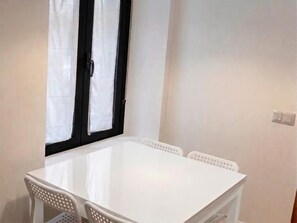 Une table blanche avec quatre chaises, une fenêtre à cadre noir et un interrupteur sur le mur.