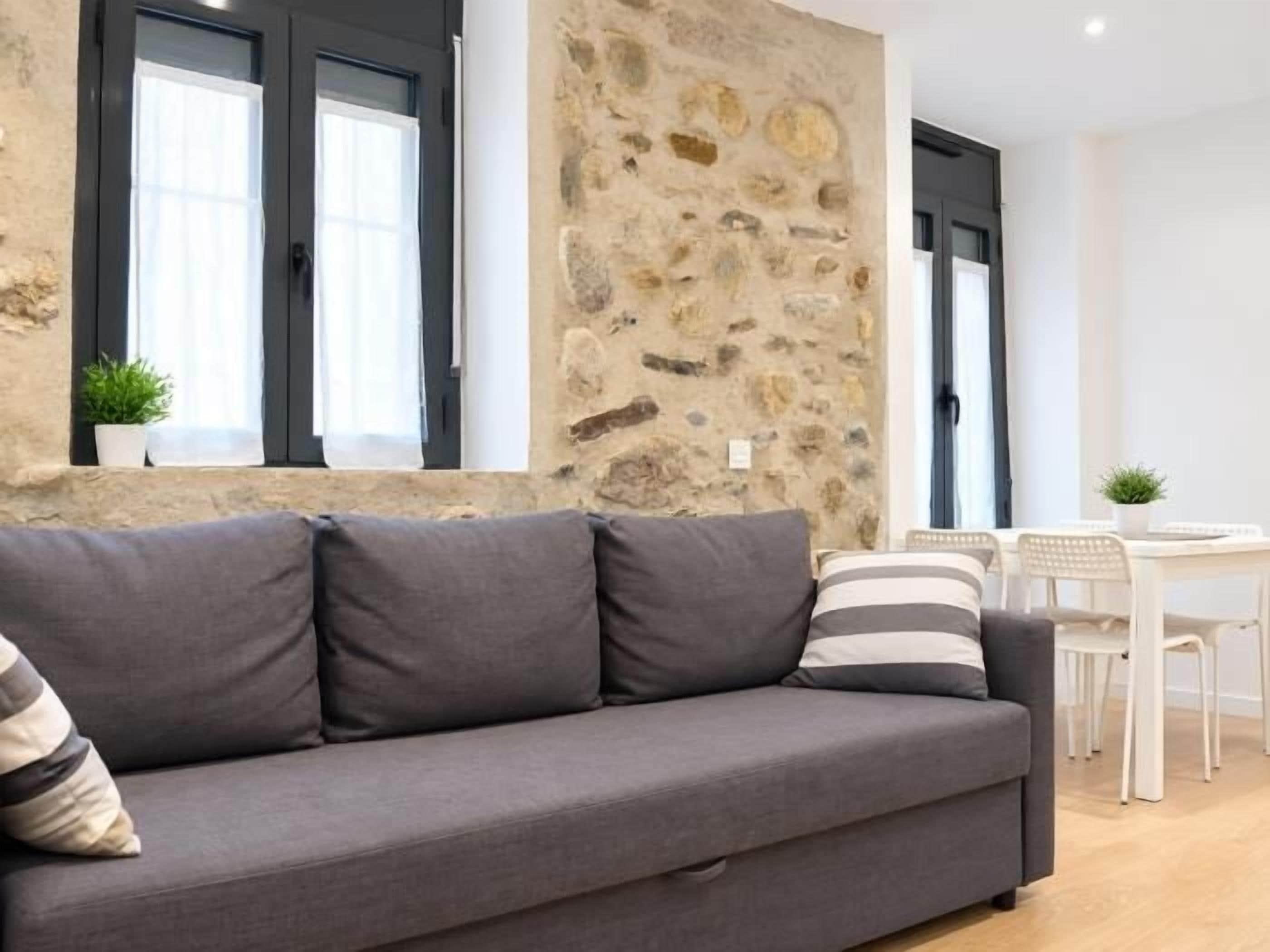 Een grijze bank met kussens, een stenen muur, een raam met gordijnen en een witte tafel met stoelen.