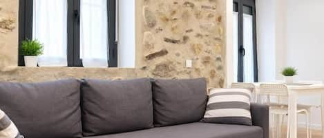 A grey sofa with pillows, a stone wall, a window with curtains, and a white table with chairs.