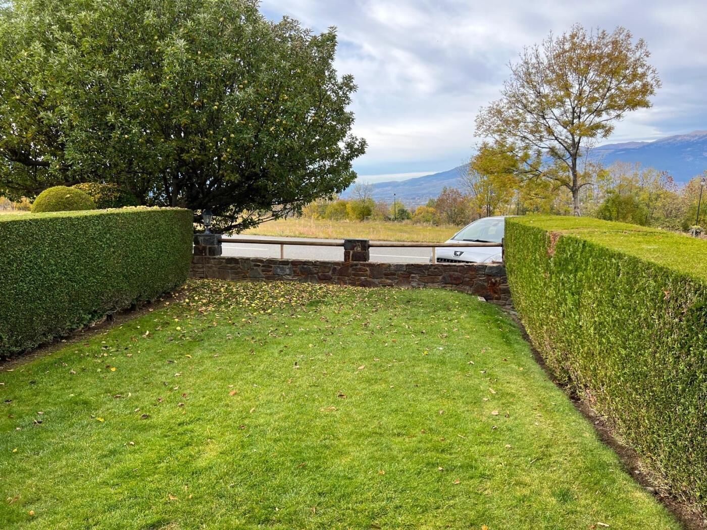 A well-maintained garden with neatly trimmed hedges, a stone wall, and a view of distant mountains.