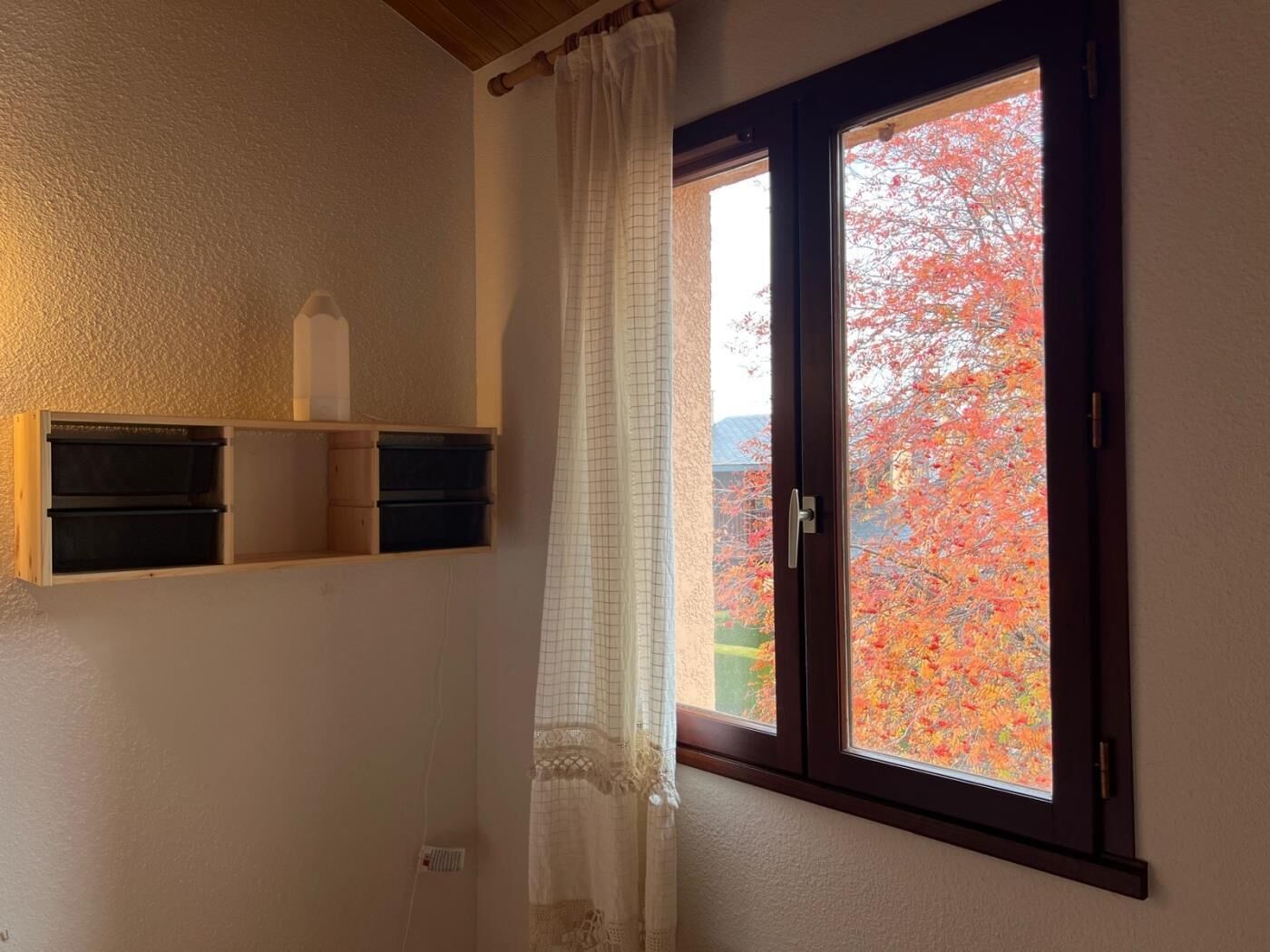 Una habitació amb finestra, una prateleira de fusta amb caixes d'emmagatzematge i una cortina blanca.