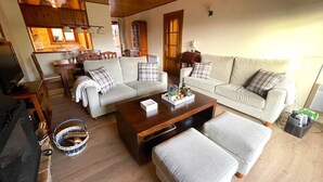 Ein gemütliches Wohnzimmer mit einer Couch, Hocker und Couchtisch, Holzboden und einem Essbereich mit Stühlen und Tisch.